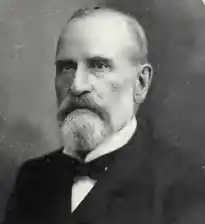Old picture of an older man with a beard, little hair, and dressed in a tuxedo