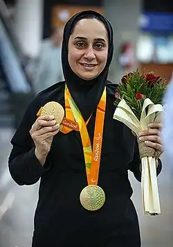 Photograpf of Javanmardi smiling; she is wearing a gold medal, holding another gold in her right hand and holding a bouquet in her left; she is dressed in black