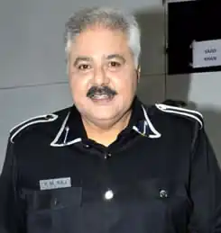 A chest up portrait of Satish Shah. He is looking at the camera and smiling. He has grey short hair and a dark moustache. He is wearing a dark shirt with white trimming on the collar and shoulders.