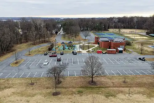 An aerial photo showing a community park. The view shows a parking lot, children playing on playground equipment, tennis courts, volleyball courts, and a baseball diamond in the distance.