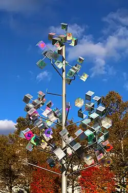 A metallic, iridescent kinetic sculpture resembling a conical tree in front of fall foliage and a blue sky.