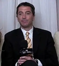 A middle-aged man sits in a chair cross-legged while staring off-screen.
