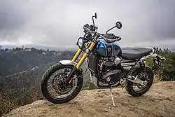 Motorcycle with a blue and black tank, long gold front suspension, and black seat. The motorcycle is parked on its side stand on a dirt road in the mountains, there are houses in the distance.
