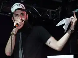 Image of the white, bearded rapper performing on stage, holding a microphone. He is wearing a black t-shirt, a grey tie and a "trucker cap". The cap has a pistol morphed into a microphone.