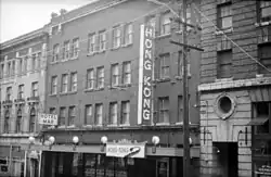 Black-and-white photo of four-story brick building with tall sign saying "HONG KONG", street-level banner saying "HONG KONG", and another sign at building's other side saying "HOTEL MAR".