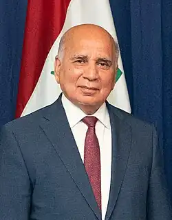 Portrait photo of a man in suit and tie against an Iraqi flag background.