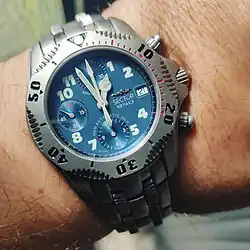 A wristwatch with a blue face and a metallic bezel, each inscribed with boldface numerals. Worn on a person's left wrist.