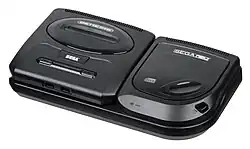Photograph of an electronic video game console resting on a compact disc attachment. The console is encased in black matte plastic that is shaped as a flattened rectangular cuboid with rounded corners. The top has two buttons and a slot for cartridges, while the front has two ports. The attachment as a top-loading disc tray with a button labeled "open".
