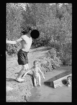 A boy swimming nude alongside another boy wearing shorts who prepares to dive into the water.