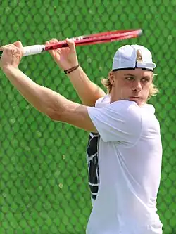 A blonde man with long hair wearing a white shirt, gray shorts, and a backwards white baseball cap holds a red tennis racket in front of his torso and above his head as he looks to his left and prepares to strike