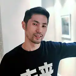 Asian man with short dark hair and light stubble smiling slightly while leaning against a wall in a softly lit room