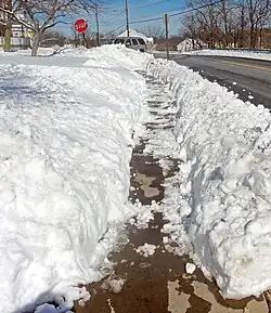 A sidewalk at the bottom of a crudely cut trench through the snow. Some snow on it is melting in the sun.