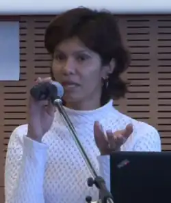 Shreen Abdul Saroor pictured giving a presentation in 2014. She stands behind a laptop and microphone stand wearing a white sweater. She is holding the microphone with one hand and is articulating with the other. Her mouth is open mid-speech.