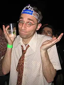 A man wearing a tiara, a loosened paisley necktie, and a pair of glasses with a beverage shrugging at the camera.