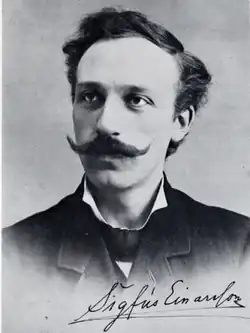 white man in 1900s costume, seated; he has wavy dark hair of medium length and substantial mustache