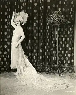 Showgirl in an ornate feathered headdress and beaded gown posing beside a patterned curtain with a long, trailing train