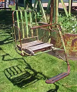 A metal chair, painted green, with a footrest and a wooden seat suspended above a grassy area by a metal pole on the right side