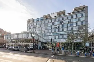 A tall concrete building seen from below and to the right, with a front facade consisting mainly of windows. Two small bare trees are in front.