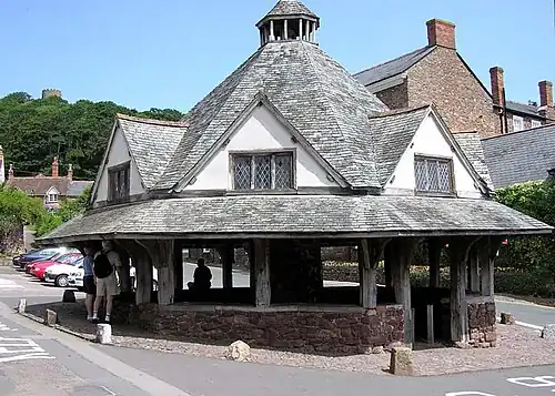 A small single-story building with a pyramid shaped roof, to the side of a road lined with buildings. Some private small cars are visible. Trees in the distance with the skyline of Dunster Castle.