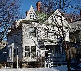 A large white wooden house with a green roof, tall chimneys, and decorative trim, partly obscured by bare trees in winter