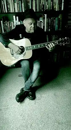Green-tinted full colour photograph of Songdog's songwriter and lead vocalist Lyndon Morgans, playing acoustic guitar at the home of Songdog's keyboard player Karl Woodward in 2025.
