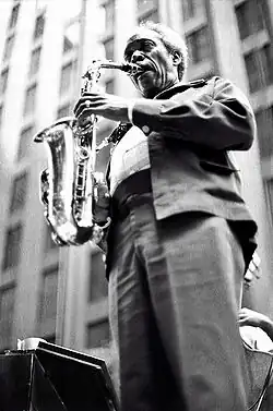 A monochrome photo of a man in a suit standing and playing a saxophone. The photo views the saxophonist from below as he plays publicly in front of a building.