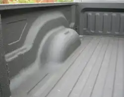 Photograph of the interior of a pickup bed covered in a gray substance. The shape of the bed is molded to allow the upper part of the wheel. There are structural ridges molded in the walls and floor.