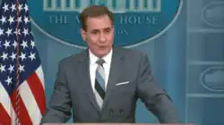 John Kirby stands at a podium in the White House briefing room, wearing a grey suit and a diagonal striped tie. He has an American flag visible to the left and a blue backdrop featuring an illustration of the White House behind him.