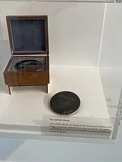 A museum display case containing a metal disc engraved 'SUFFOLK' beside a wooden presentation box. A label reads: 'The Suffolk Medal was designed by renowned Suffolk artist, Maggi Hambling, reflecting her view of Suffolk. The medal is contained in a box made of Suffolk walnut, personalised for the recipient.'