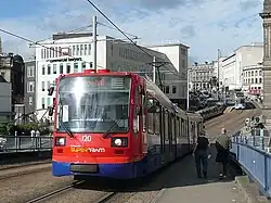 A Sheffield Supertram in current blue, orange and red Stagecoach livery. The tram shown is crossing Park Square bridge and Fitzalan Square and Castle Square can be seen in background, as can tram tracks and numerous commercial buildings.