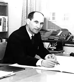 Black and white photograph of a man wearing a suit and sitting at a desk. He has a pen in his hand, and there is a telephone and dictaphone on his left and a bookshelf behind him