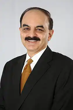 A man in a black suit, white shirt, and tan tie is smiling at the camera. He has a black moustache. The background is light gray.