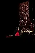 A female opera singer, located in the center of the image on-stage, sings the national anthem of Draag. A screen showing the lyrics to the song as well as background footage themed around Draag is shown behind her.