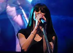 Sylwia Grzeszczak singing into a microphone on stage during a concert in Wrocław. She is/has holding her microphone against a blue-purple background.