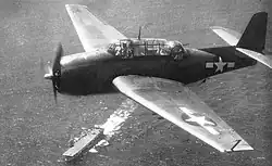 A propellered aircraft in Navy livery flying above the ocean. In the background is an aircraft carrier. The number seventy-seven is painted onto the front of the aircraft carrier's flight deck. At the end of the flight deck, another plane prepares for take-off.
