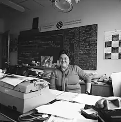 black-and-white image of Tatiana Fabergé grinning at camera from behind a desk covered in papers and office instruments, blackboard covered in writing in background
