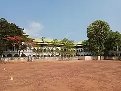 A two-story educational or institutional building with distinctive Islamic architecture featuring multiple arched doorways and windows across both levels. The structure has a light green or yellow-green roof and white walls with decorative arches. A low wall with balustrades runs along the front of the building. The foreground shows a large red dirt sports ground or playground with cone markers. Mature trees, including one with reddish-orange foliage, frame the building on both sides against a clear blue sky.
