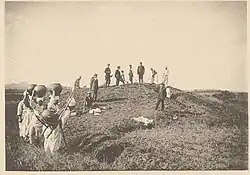 A sepia-toned photograph of a group of archaeologists standing on a tumulus. A group of people in white caps stand in the foreground, carrying large vases above their head
