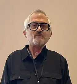 Picture of person on a beige background, wearing thick black glasses