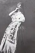 A black-and-white photograph of a woman dressed as a tapada limeña, holding a shawl embroidered with floral patterns and partially covering her face with a dark veil.