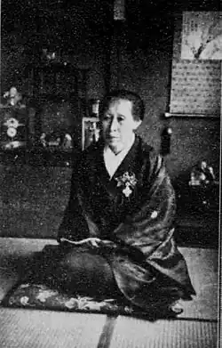 An older Japanese woman, seated on a floor cushio, wearing a kimono