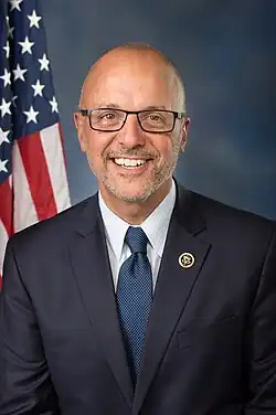 A chest-up photo of bald man wearing glasses and a suit, in front of a blue background