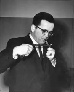 Ted Stevens lighting a pipe on January 23rd, 1967. He is in a dark room, wearing glasses, a black suit, and a black tie. His head is tilted downwards, and his body is tilted slightly to the right hand side of the photograph.