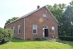 A small red‑brick church with white trim, rectangular in form, featuring a central entrance and symmetrical windows, surrounded by grass and trees.