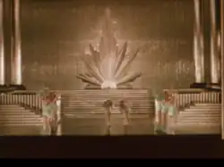 A group of dancers performs on a grand Art Deco stage with tiered steps and a radiant sunburst backdrop.