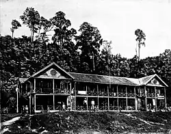 Big friggin wood building in jungle, old photo, black and white. Palm trees in background. Couple of guys posted up front chillin. Imagine the heat of malaria and then imagine working in khakis.