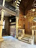 This ornate hall features a ceiling richly decorated with golden geometric and floral patterns. The space is framed by elegant, rhythmically placed arches in bold black-and-white stripes. Hanging lamps illuminate the interior, creating a sacred and timeless atmosphere.