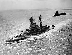 A large warship steaming through a calm sea with a large, flat-decked warship following behind