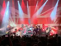 Shot of a concert stage. A large Union Jack flag backs the stage with musical equipment and four band members lit in white and red lighting in front of it. At extreme left is a male playing guitar. Central stage has a lead vocalist singing and a drummer and his kit to his right. Right stage has a bass guitarist.