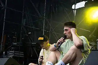Two men sit cross-legged on a music festival stage, holding microphones. Both are wearing shorts and a T-shirt, the one on the left is wearing a black baseball cap featuring the Pirelli logo. A bright stage light flares in the background.